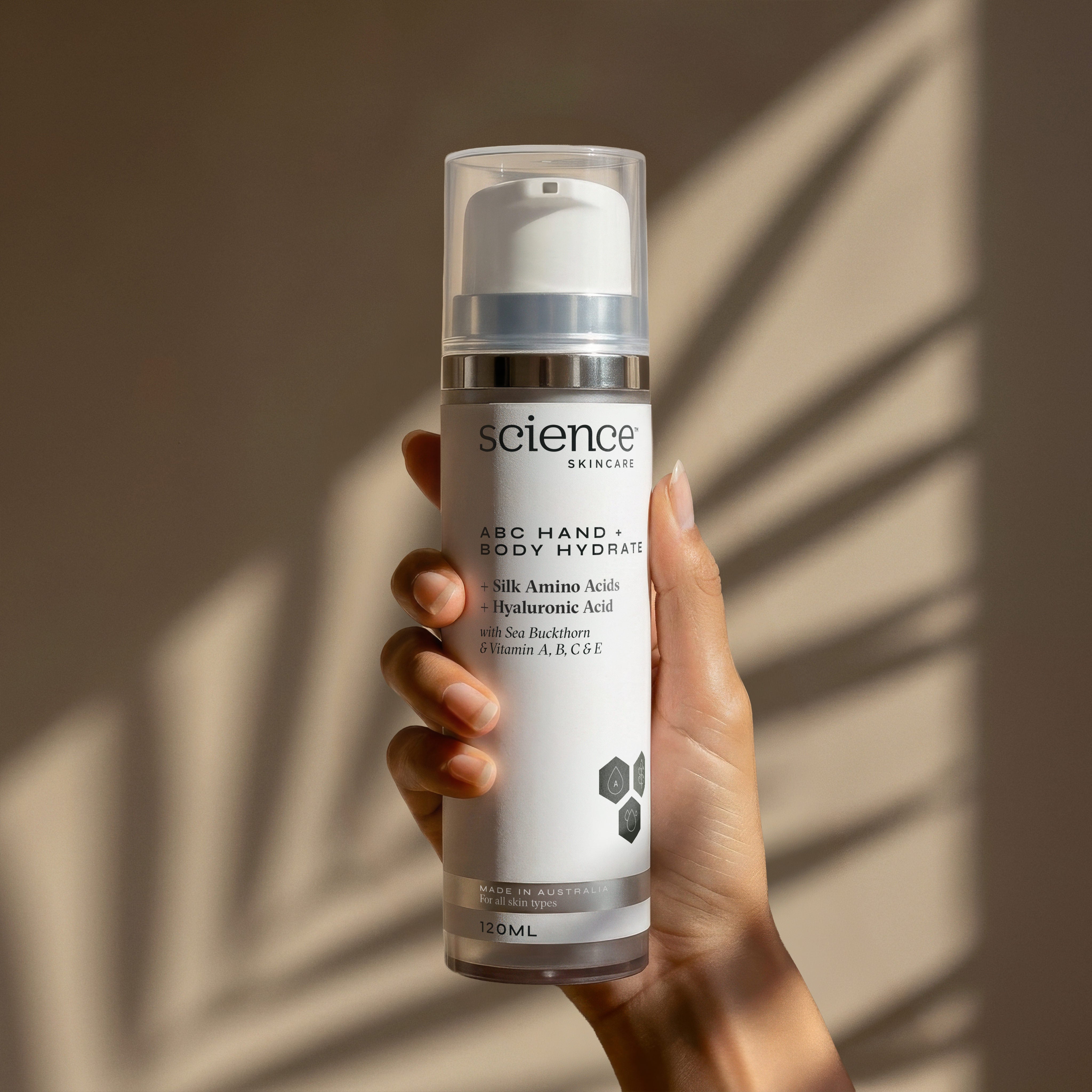 ABC Hand + Body Hydrate lifestyle image showing a hand holding the 120ml Science Skincare body moisturiser with silk amino acids, hyaluronic acid, sea buckthorn and vitamins A, B, C and E, against a warm wall with dappled palm shadow play
