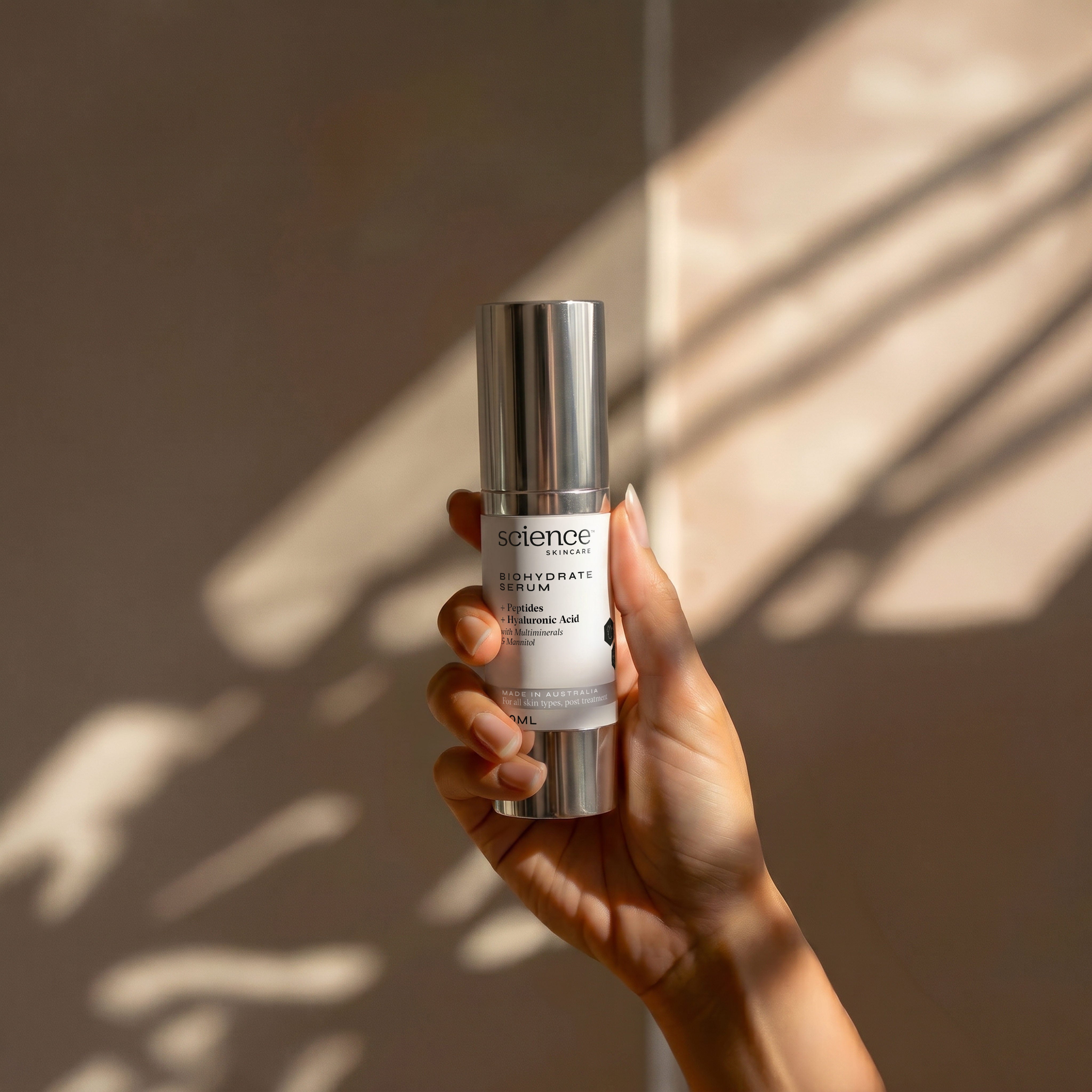 Biohydrate Serum lifestyle image showing a hand holding the 30ml Science Skincare hydrating serum with peptides, hyaluronic acid, multiminerals and mannitol, against a warm wall with dappled light shadows
