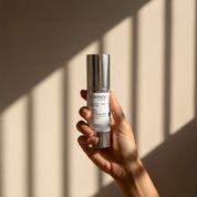 Clear + Calm Serum lifestyle image showing a hand holding the 30ml Science Skincare serum with retinyl, alpha and beta-hydroxy acids, Australian tea tree oil and manuka honey, against a warm wall with geometric light shadows
