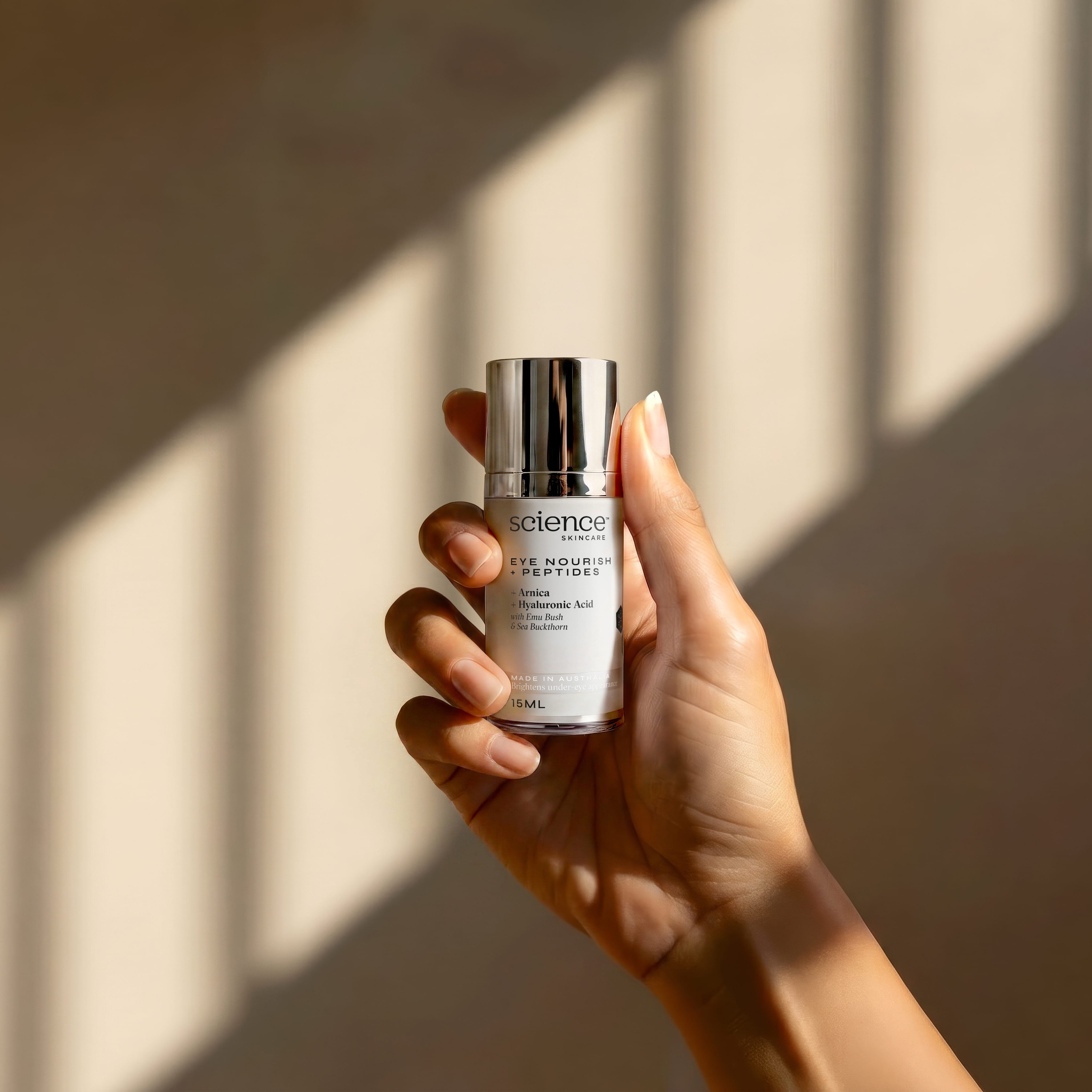 Eye Nourish + Peptides lifestyle image showing a hand holding the 15ml Science Skincare eye treatment with arnica, hyaluronic acid, emu bush and sea buckthorn, against a warm wall with geometric light shadows