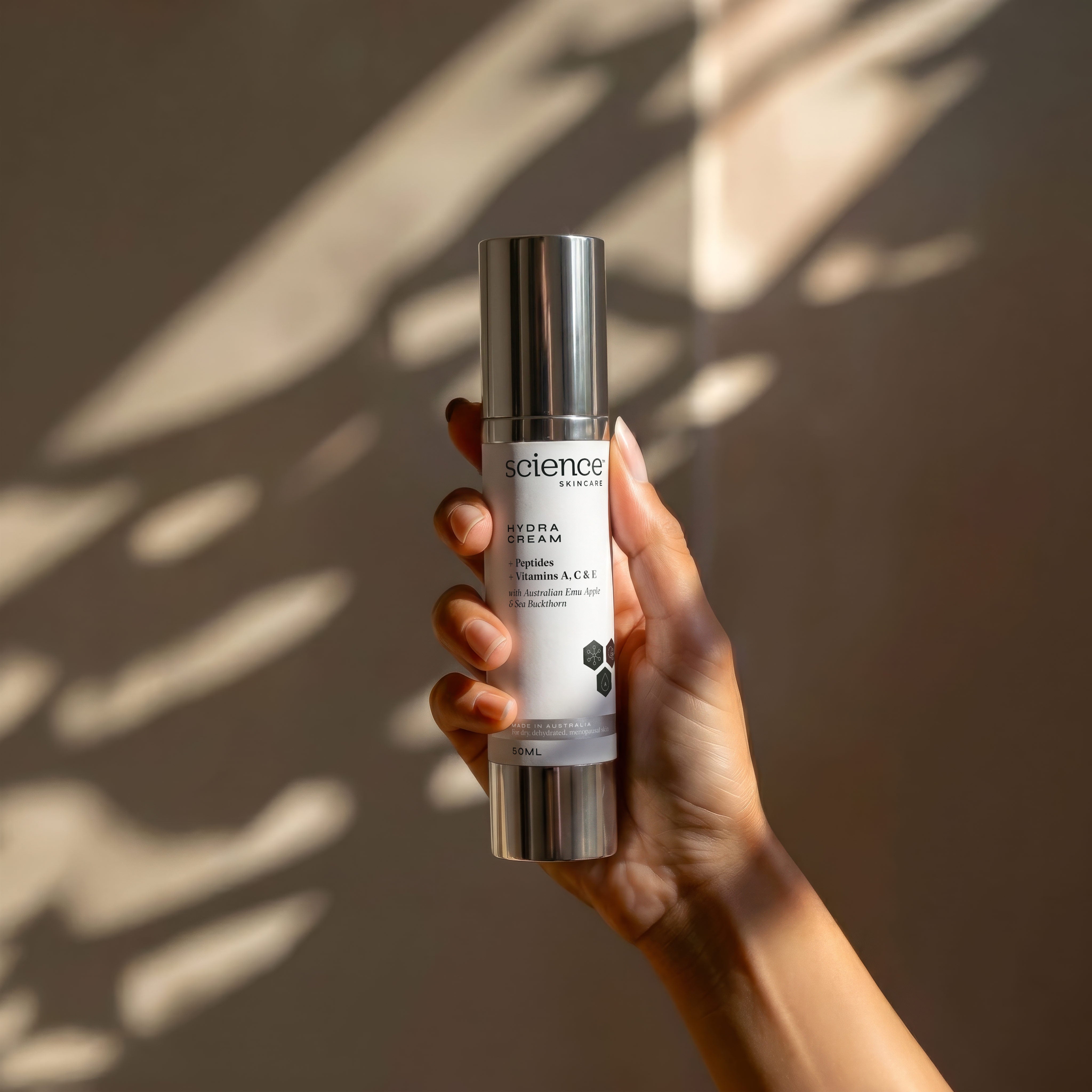 Hydra Cream lifestyle image showing a hand holding the 50ml Science Skincare face moisturiser with peptides, vitamins A, C and E, Australian emu apple and sea buckthorn, against a warm wall with dappled light shadows