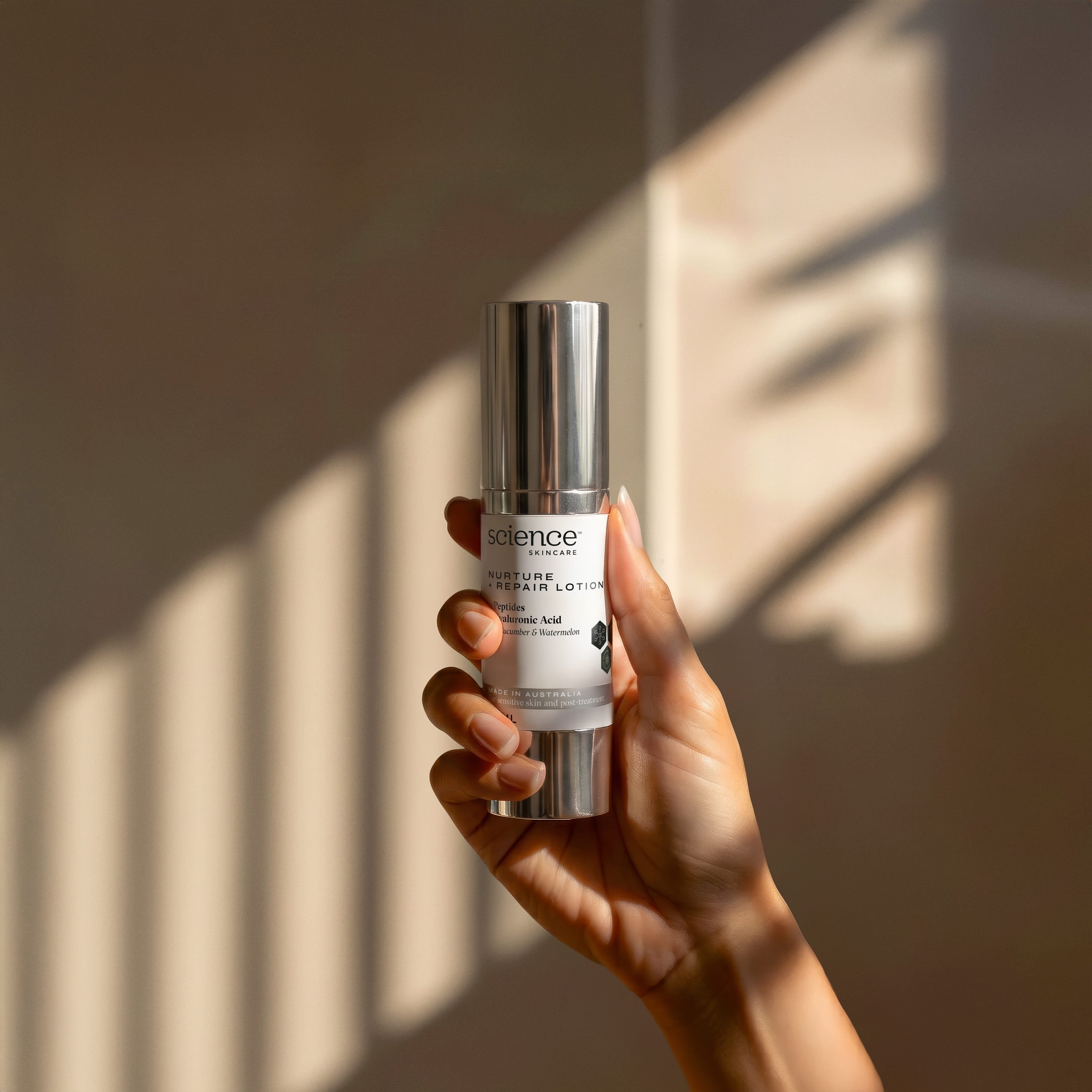 Nurture + Repair Lotion lifestyle image showing a hand holding the 30ml Science Skincare soothing lotion with peptides, hyaluronic acid, cucumber and watermelon, against a warm wall with geometric light shadows