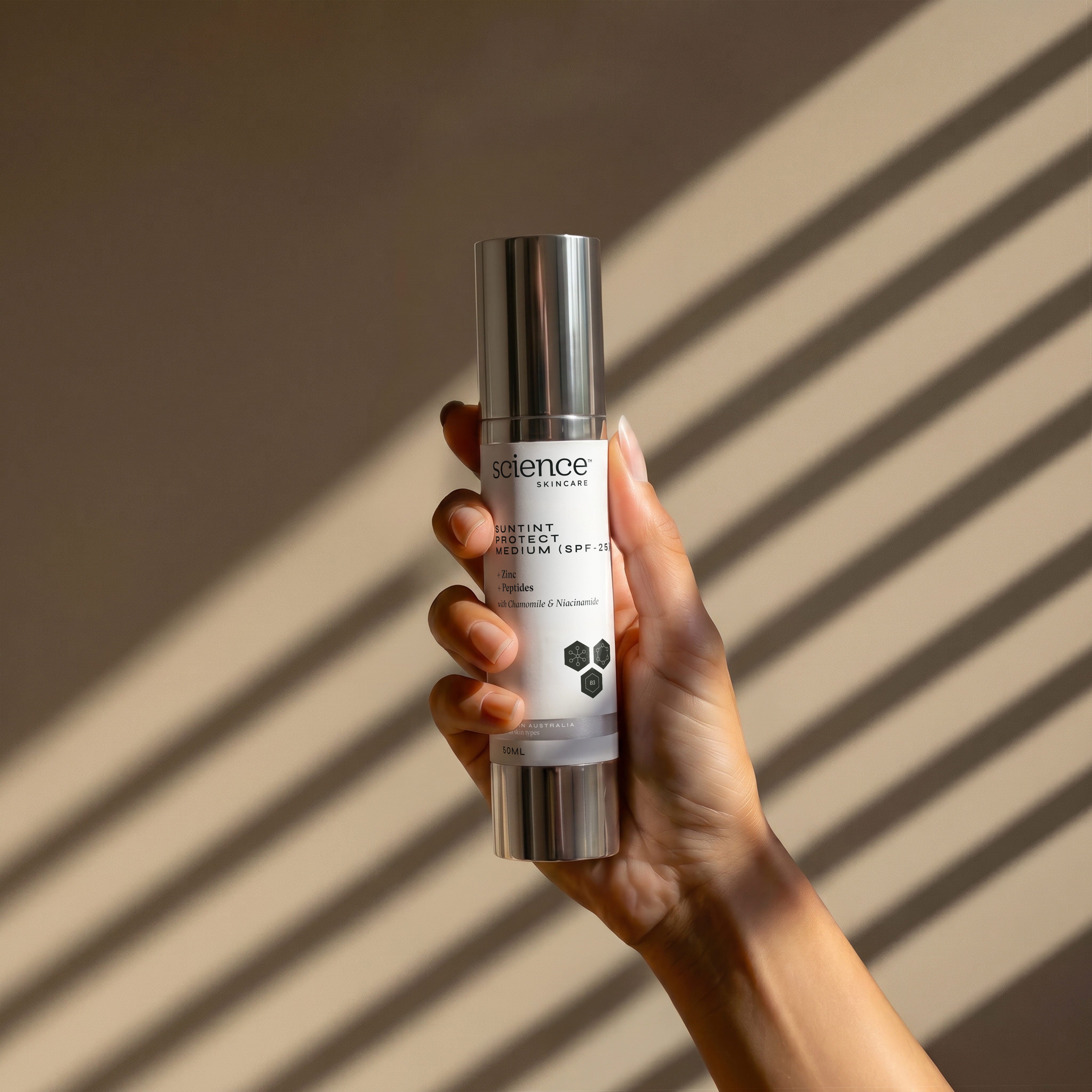 Suntint Protect Medium SPF-25 lifestyle image showing a hand holding the 50ml Science Skincare tinted sunscreen with zinc, peptides, chamomile and niacinamide, against a warm wall with diagonal light shadows

