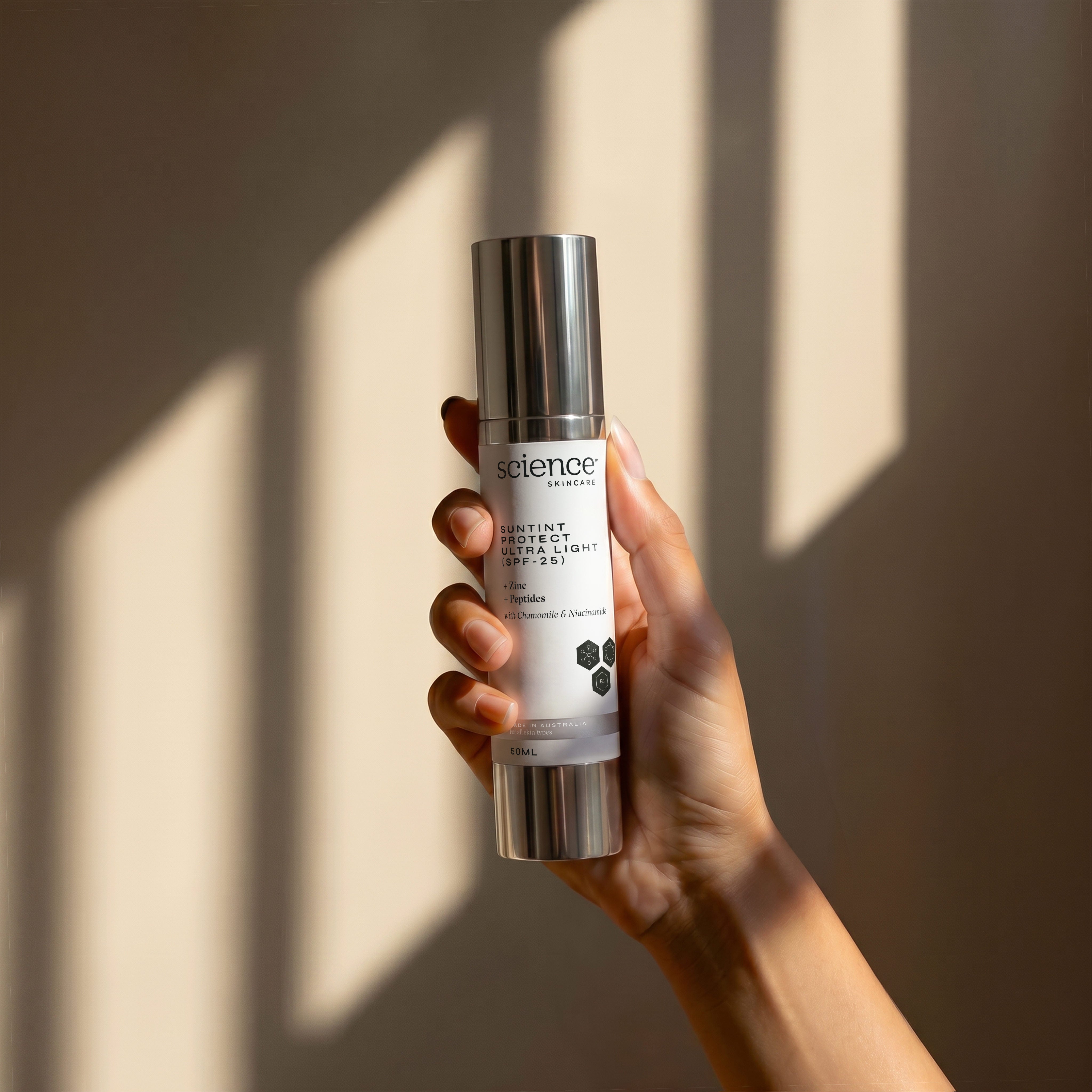 Suntint Protect Ultra Light SPF-25 lifestyle image showing a hand holding the 50ml Science Skincare tinted sunscreen with zinc, peptides, chamomile and niacinamide, against a warm wall with geometric light shadows
