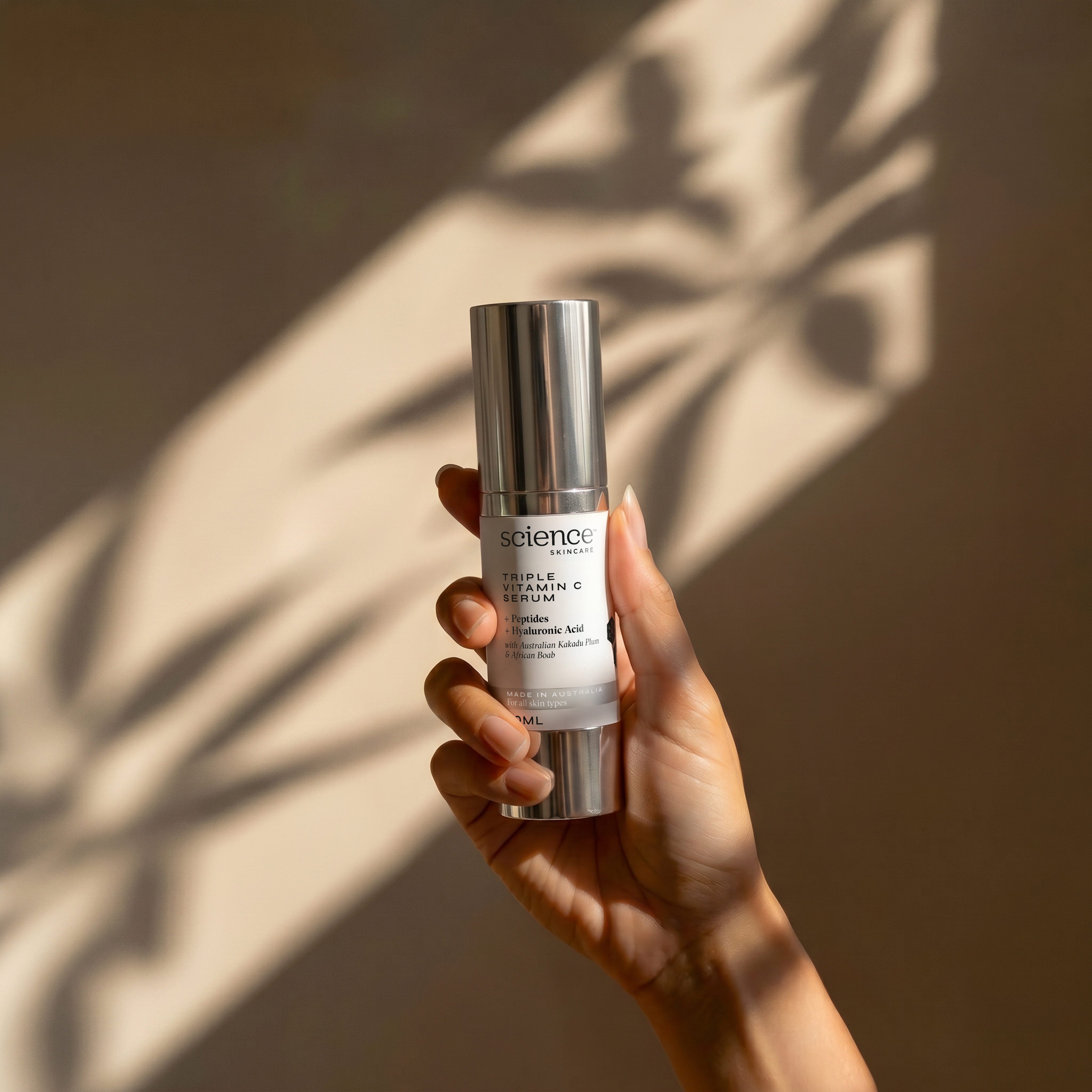 Triple Vitamin C Serum lifestyle image showing a hand holding the 30ml Science Skincare vitamin C serum with peptides, hyaluronic acid, Australian kakadu plum and African baobab, against a warm wall with dappled palm shadow play
